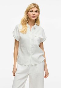 Camicia bianca a maniche corte con chiusura a bottoni, una tasca sul petto e un tessuto morbido e testurizzato. Indossata con pantaloni bianchi coordinati.