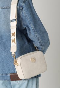 Vit texturerad axelväska med en gyllen logotyp, silverdetaljer och ett vitt band med guldaccenter. Bärs över en jeansjacka.
