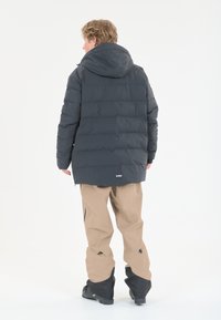 Dunkelgraue Pufferjacke mit Kapuze, gesteppter Textur und Logodetail am Saum; kombiniert mit beigen Hosen mit Reißverschlüssen an den Knien.
