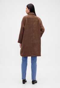 Casaco comprido xadrez castanho com gola larga, corte oversized e tecido texturizado, combinado com jeans azul claro e botas pretas.