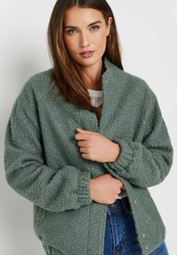 Green, textured, oversized jacket made of soft fabric with a high collar and snap button closure. The sleeves are gathered at the cuffs.