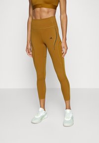 Leggings marrones de talle alto con costuras negras en contraste, bolsillos con cremallera y una cinturilla elástica; combinados con zapatillas de deporte verde claro.