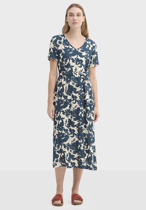 Robe mi-longue à motifs floraux en bleu foncé et crème, avec manches courtes, col en V et ceinture en tissu. Portée avec des sandales rouges.