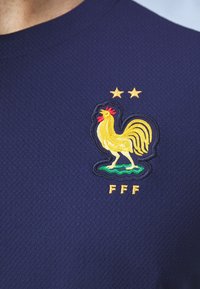 Námořnická modrá texturovaná sportovní košile se žlutým vyšitým logem kohouta, dvěma hvězdami nad ním a zlatým nápisem "FFF" pod ním.