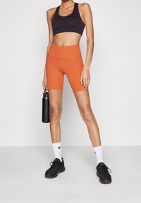 Černá sportovní podprsenka, oranžové šortky s vysokým páskem, černé tenisky, bílé ponožky s logem. Lahvička s vodou držena v pravé ruce. Hladká textilní struktura.