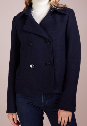 Femme portant un manteau en laine bleu marine à double boutonnage par-dessus un col roulé blanc et un jean bleu foncé, debout devant un fond uni.