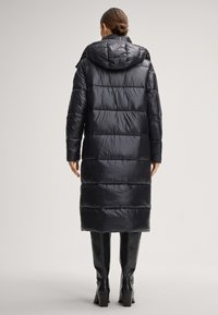 Długa czarna pikowana kurtka puchowa z kapturem, poziomy wzór pikowania, błyszcząca nylonowa tekstura, sięgająca do połowy łydki, noszona z czarnymi butami za kolano.