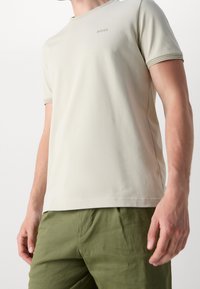T-shirt a maniche corte color beige chiaro con scollatura tonda, caratterizzato da sottili accenti verdi sul colletto e sulle maniche, realizzato in materiale morbido.
