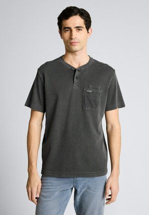 Jonge man met kort donker haar die een donkergrijs henley T-shirt met korte mouwen en een borstzak draagt, evenals lichtblauwe spijkerbroek.