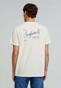 Creme bomulds t-shirt med løs pasform, der har et iøjnefaldende blåt grafisk print på ryggen, der siger "Scalpers Co" og hotelinformationer.