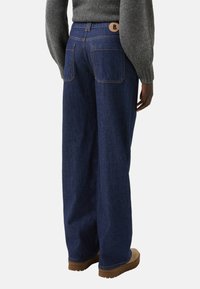 Jeans w stylu wide-leg w ciemnoniebieskim kolorze, z wysokim stanem i logo na guziku, tylne kieszenie oraz luźny krój. Materiał bawełniany o teksturze.