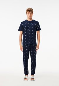 Navyblauw pyjama set met een T-shirt met korte mouwen en lange broek met witte vissenvormen; gecombineerd met witte sandalen met groene accenten.