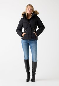 Zwarte pufferjack met een met bont gevoerde capuchon, gouden hardware, verstelbare taille met riemen, gecombineerd met strakke blauwe jeans en zwarte kniehoge laarzen.