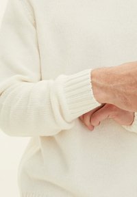 Cremfarbener Strickpullover mit gerippten Bündchen, der eine weiche Textur und einen lockeren Schnitt präsentiert. Hände sind sichtbar, eine hält das Handgelenk der anderen.