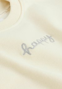 Cremefarvet sweatshirt med ribbet halsudskæring, prydet med sølvbroderet tekst "happy" på forsiden. Blød tekstur, afslappet design.