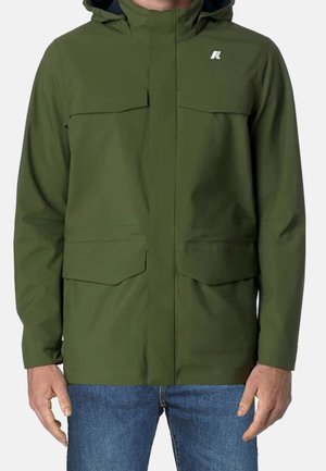 Homme portant une veste à capuche vert olive avec des poches avant et un jean bleu, debout devant un fond uni.