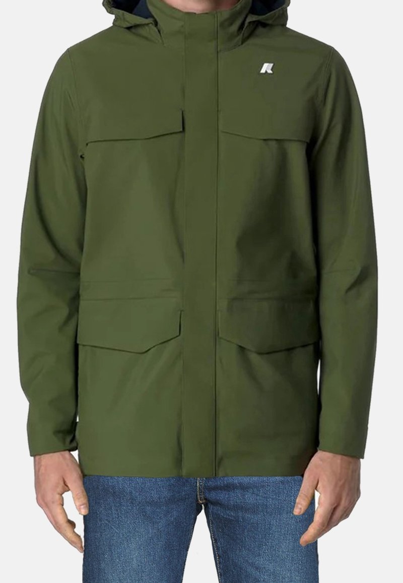 Homme portant une veste à capuche vert olive avec des poches avant et un jean bleu, debout devant un fond uni.