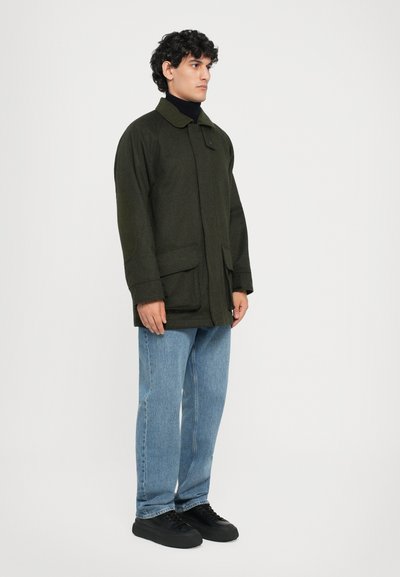 Olive green jacket with a collar, two front pockets, and a relaxed fit, paired with light blue jeans and black sneakers.