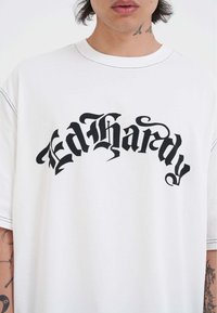 Person med en hvid t-shirt, der har sort gotisk tekst "FedHardy" over brystet, synlig hals og delvise tatoveringer på armene.
