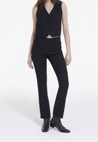 Schwarze, gestreifte Weste und passende Hose, beide tailliert. Die Weste hat einen V-Ausschnitt und Knöpfe, während die Hose gerade geschnitten und mit einer glatten Oberfläche versehen ist.