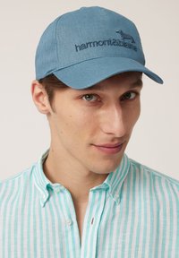 Blue cap with embroidered logo and dog design, made of textured fabric. Worn with a light blue and white striped button-up shirt.