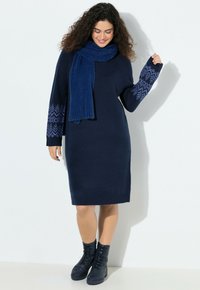 Abito a maglione blu navy con maniche decorate, polsini a coste e una sciarpa blu abbinata. Completato da stivali neri con lacci alla caviglia.