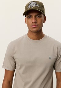 Μπεζ κοντομάνικο μπλουζάκι με στρογγυλή λαιμόκοψη. Διαθέτει ένα μικρό πράσινο λογότυπο "II" στο αριστερό μέρος. Ελιά καπέλο με λευκό γραφικό σχέδιο.