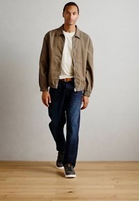 Helepruun bomber-jakk, elastsete mansettidega, valge T-särk, tumedast sinisest denimsest teksad ja hallid spordijalatsid. Puitpõranda taust.