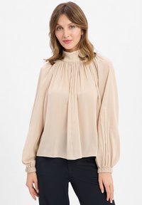 Blusa beige realizzata in tessuto leggero con maniche lunghe a sbuffo e collo alto. Presenta dettagli arricciati lungo il petto.