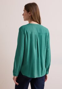 Blusa verde a maniche lunghe in tessuto morbido con taglio dritto, piega posteriore e orlo arrotondato. Polsini con bottoni e dettagli minimi.