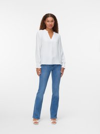 Hvit bluse med lange ermer, V-hals og teksturert stoff, kombinert med lyseblå flagrende jeans og hvite sandaler med åpen tå.