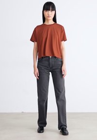 Vrouw die staat met een roestkleurig shirt met korte mouwen, hoge zwarte jeans en zwarte schoenen voor een effen witte achtergrond.