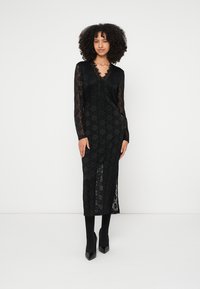 Svart spetsklänning med långa ärmar, V-ringning, blommönster, högt slits på låret, kombinerad med svarta strumpbyxor och spetsiga klackar.