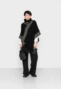 Poncho noir avec accent argenté, ourlet effiloché, col haut, porté sur une chemise blanche, associé à un pantalon noir à jambes larges et un sac à main texturé.