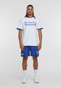 Hvit oversized T-skjorte med blå kant og teksten "New York City Basketball", kombinert med blå mesh-shorts, hvite sokker og joggesko.