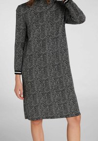 Robe en tricot en chevron noir et blanc, longueur genou, avec des manches longues et un col haut. Présente des poignets rayés aux extrémités des manches.