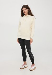 Felpa crema con tasca a marsupio, abbinata a leggings neri con testo grafico bianco e scarpe bianche con lacci rosa.
