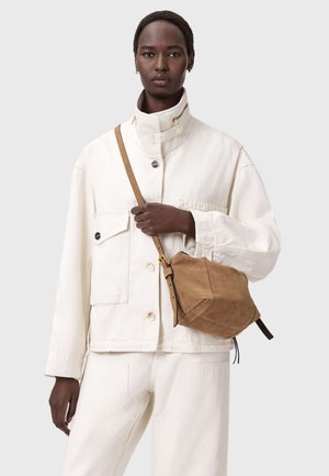 Borsa a tracolla in camoscio marrone a forma triangolare, con texture morbida e tracolla regolabile, indossata con giacca e pantaloni bianchi.