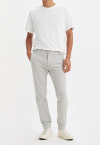 Man wearing a plain white T-shirt, light gray pants, and off-white high-top sneakers, standing with hands in pockets.