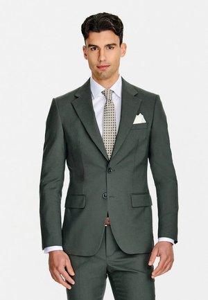 Mężczyzna w ciemnozielonym garniturze, białej koszuli, wzorzystym krawacie i białej poszetce, stojący na jednolitym białym tle.