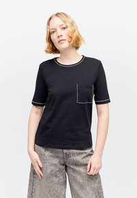 Jeune personne aux cheveux blonds portant un t-shirt noir à manches courtes avec des coutures blanches et un jean gris délavé à jambes larges.