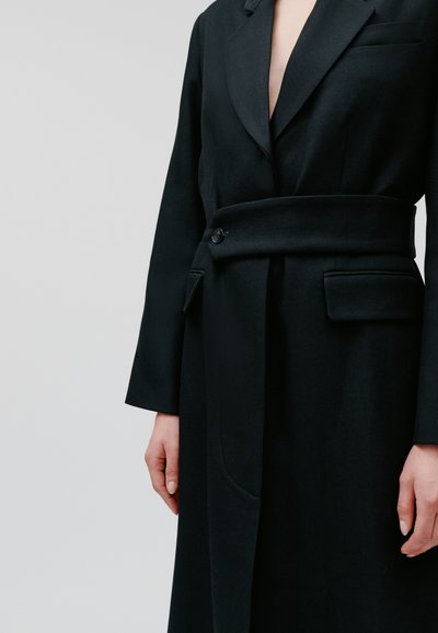 Persona che indossa un lungo cappotto nero su misura con una cintura larga fissata da un bottone in vita, in piedi contro uno sfondo chiaro e semplice.