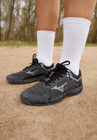 Scarpe sportive in nero e grigio, con una tomaia testurizzata, suola robusta e accenti riflettenti. Abbinati a calzini bianchi a coste.