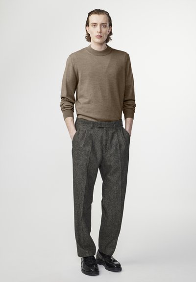 Bruine gebreide trui met een hoge halslijn, gecombineerd met grijze tweed broek met plooien en zwarte leren schoenen. Eenvoudig ontwerp, op maat gesneden.