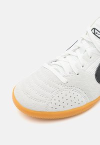 Zapatilla de ante blanca con puntera perforada, cordones blancos, logo negro y suela de goma naranja sobre un fondo blanco.