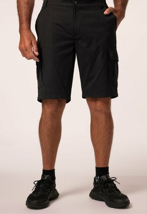 Mand iført sorte cargoshorts, sorte sokker og sorte sportssko står med den ene hånd i lommen.