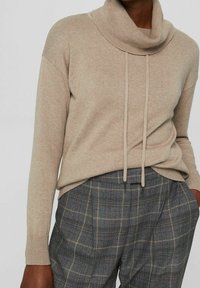 Pull beige en maille avec col bénitier et cordons, manches longues. Associé à un pantalon taille haute à carreaux gris.