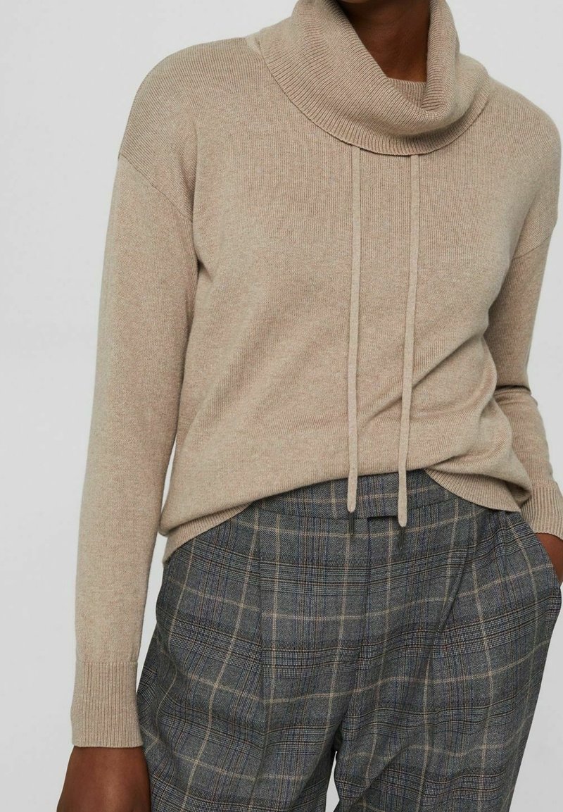 Pull beige en maille avec col bénitier et cordons, manches longues. Associé à un pantalon taille haute à carreaux gris.