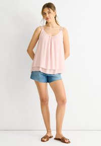 Top senza maniche di un delicato rosa chiaro con decorazioni floreali sulle spalline, design a strati, abbinato a pantaloni corti in denim casual e sandali marroni.