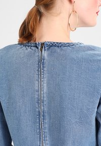 Denimmekko, jossa on korkea pääntie, takana vetoketjukiinnitys ja punottu tikkaus kauluksen ympärillä. Vaalea sininen kangas, jossa on pehmeä tekstuuri.